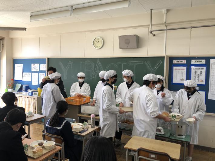 給食を準備している白衣の学生。黒板にはyuba cityの文字がある。