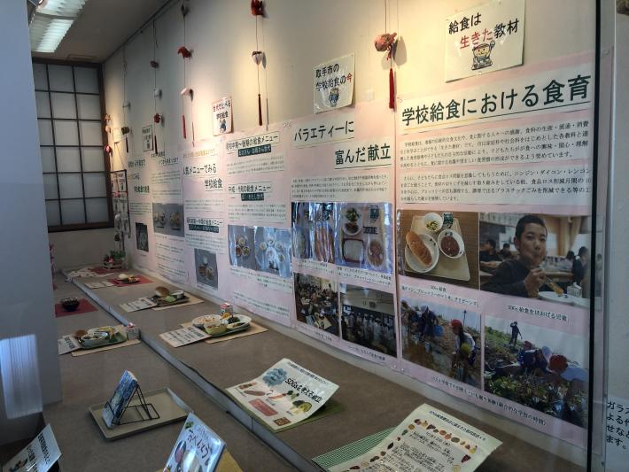 壁面の展示スペースには、給食を食べている子どもの写真や昔の給食が展示されている。床には、本や給食のサンプルが置かれている。