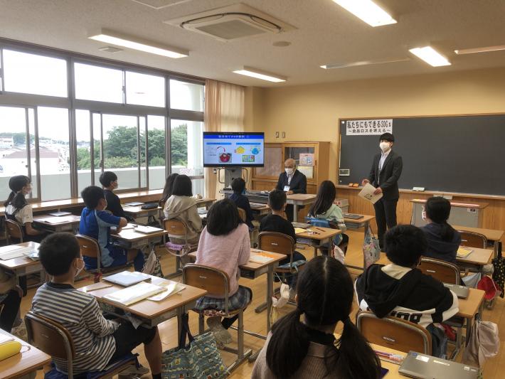 大型掲示装置に映されたスライドの説明をする職員とそれを座って聞く児童