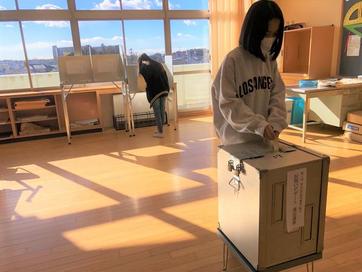 投票箱に投票用紙を投函する女子児童。その後ろにある投票記載台で児童1名が候補デザートを選んでいる。