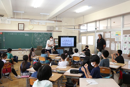 杉浦先生が児童に演台付近のモニター前でSDGsの活動例を提案している様子