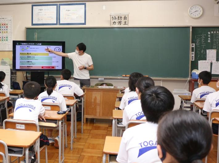 杉浦先生が教室の黒板脇に設置されているモニターを用いて参加している生徒に説明している様子