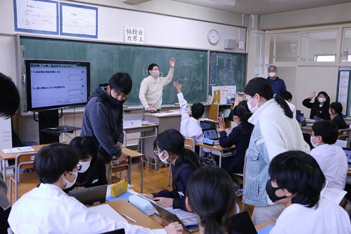 教室で先生が教壇に立ち、生徒に挙手を求め、生徒が挙手等をしている