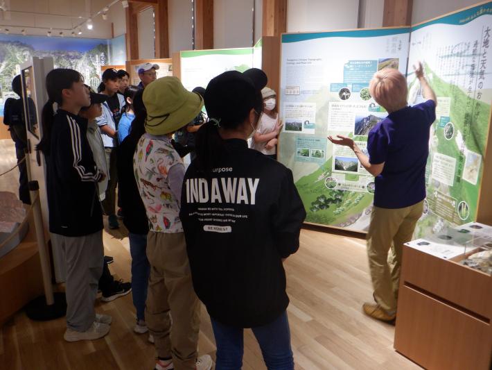 谷川岳インフォメーションセンターで、高山植物の案内板の前で説明をする自然解説ガイドとその周りで話を聞く児童たち