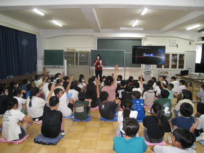 児童たちが教室で座って話を聞いている。何人かの児童は手を上げている