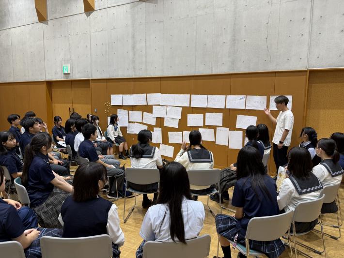 いろいろな内容が書かれたA4の紙がたくさん貼られた前で一人が発表している。その前には椅子に座ったその他の高校生たち。
