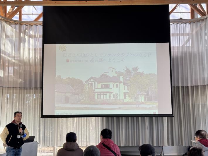 スクリーンの前で男性がマイクを持ち挨拶している様子