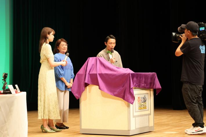 一般の依頼人がステージ上でお宝について司会と話している様子