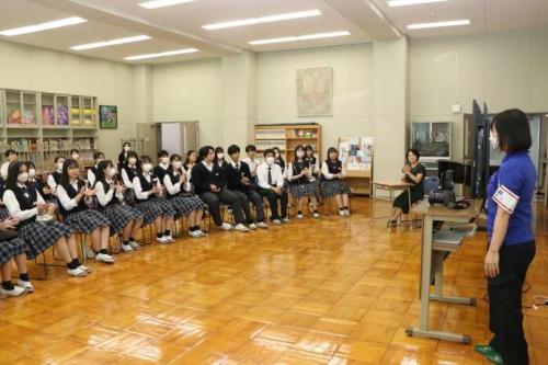 椅子を並べて並んで座っている生徒たちの前で、若い女性が何かを話しかけている。