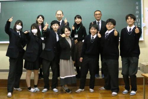 学生と先生、若い女性の記念写真。黒板前に2列に並んだ人たちが親指を前に出してポーズしている