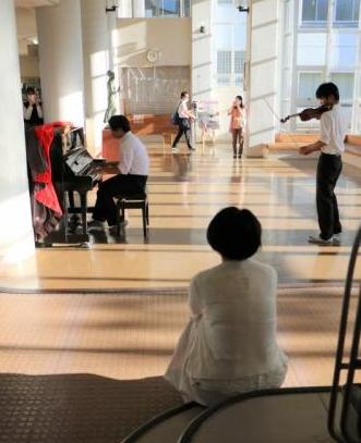ホールでのピアノとバイオリン演奏。それを見守る若い女性の後ろ姿の写真。