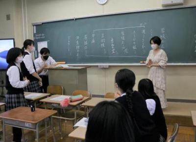 黒板に10程度の言葉が羅列して書き出している若い短髪女性とそれを見ている生徒の写真。