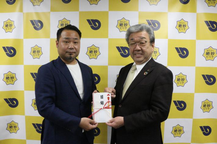 黄色と白のパネルの前で、左側に鈴木代表、右側に市長が並んで立っている。