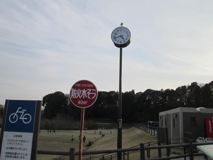 高井小学校側駐車場脇に設置された時計。隣には防火水槽や駐輪場の標識が並んでいる。