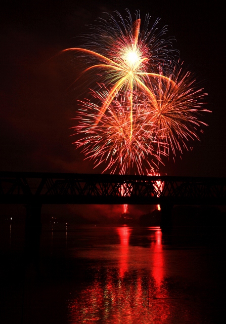 打ち上げられた花火。夜空に浮かび上がる鉄橋を背景に赤い色の尺玉数発が打ち上げられ開いている画像。