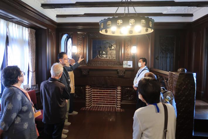 旧渡辺甚吉邸内の洋間で、装飾の施された照明や暖炉についてガイドの説明を受ける参加者の様子