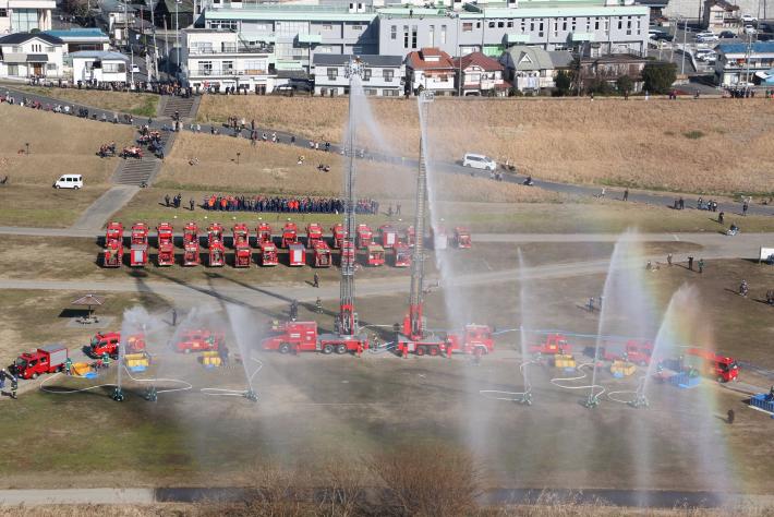 消防出初式の様子。河川敷に消防車が並び、放水訓練を行っているところを上空から撮影した写真