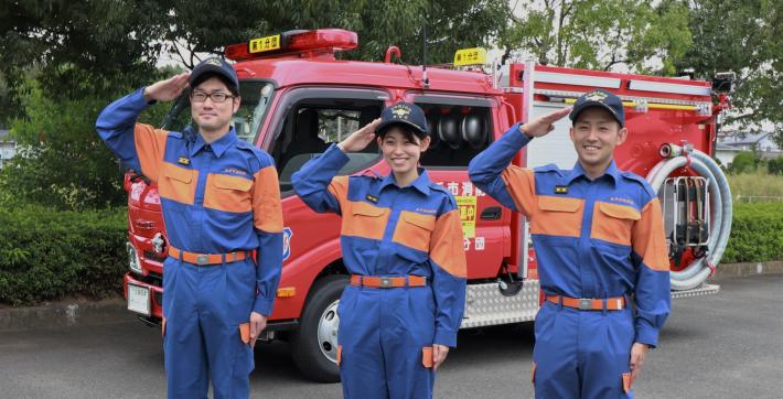 消防車両を背景に3人の団員が敬礼をしている写真。