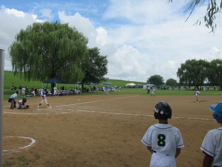 子どもたちが野球をしています。グラウンドの手前で8の背番号のユニフォームの子どもが遠くの試合の様子を見ている画像。
