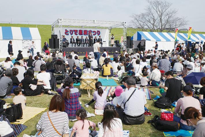 とりで産業まつり。ステージであいさつをする人、後ろでたくさんの人たちが座りながら聞いている。